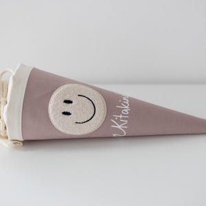 May include: A dusty rose fabric cone-shaped gift bag featuring a cream-coloured circular patch with a black embroidered smiley face. The top of the bag has a cream fabric collar with a drawstring closure and the white text Kitaking printed on the side.