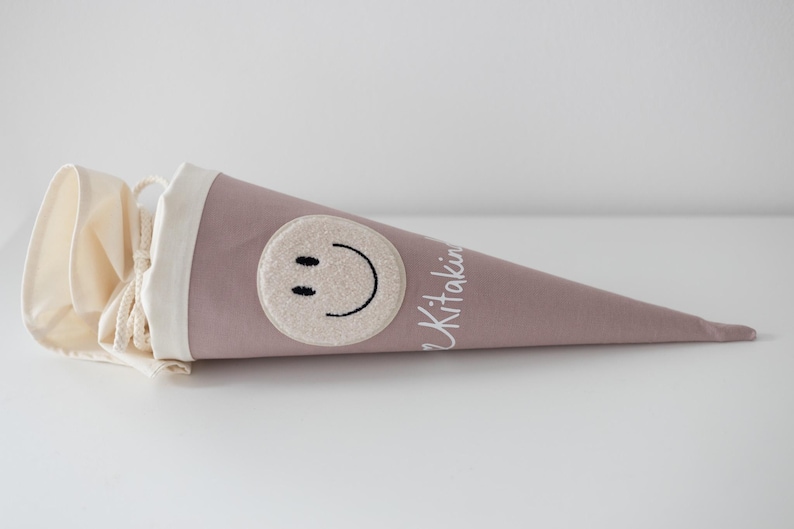 May include: A dusty rose fabric cone-shaped gift bag featuring a cream-coloured circular patch with a black embroidered smiley face. The top of the bag has a cream fabric collar with a drawstring closure and the white text Kitaking printed on the side.