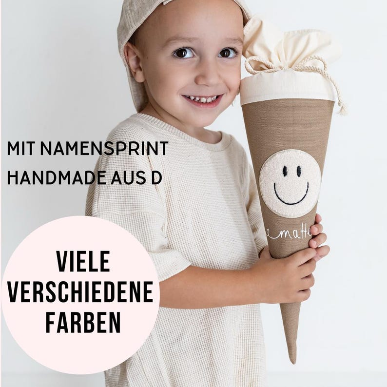 May include: A beige fabric cone-shaped gift bag featuring a circular patch with a black embroidered smiley face, a cream-coloured drawstring top, and the name matte written in white script. Text reads: MIT NAMENSPRINT HANDMADE AUS D VIELE VERSCHIEDENE FARBEN.