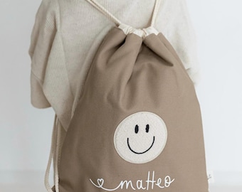 May include: A taupe drawstring backpack featuring a circular cream-coloured patch with a black embroidered smiley face. Below the patch, the name "matteo" is embroidered in white cursive script, accompanied by a small heart icon on the left side.