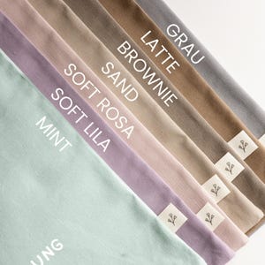 May include: A collection of fabric pouches arranged in a diagonal fan pattern, featuring colours labelled as Mint, Soft Lila, Soft Rosa, Sand, Brownie, Latte, and Grau. Each pouch includes a small white fabric tag with a simple black floral icon.