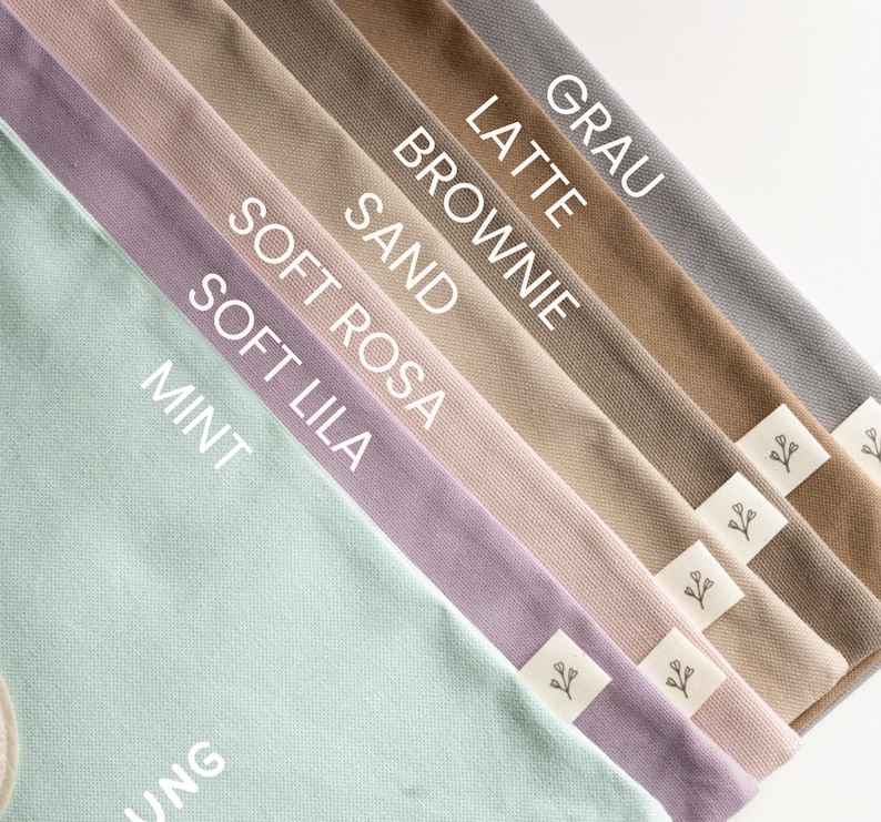 May include: A collection of fabric pouches arranged in a diagonal fan pattern, featuring colours labelled as Mint, Soft Lila, Soft Rosa, Sand, Brownie, Latte, and Grau. Each pouch includes a small white fabric tag with a simple black floral icon.