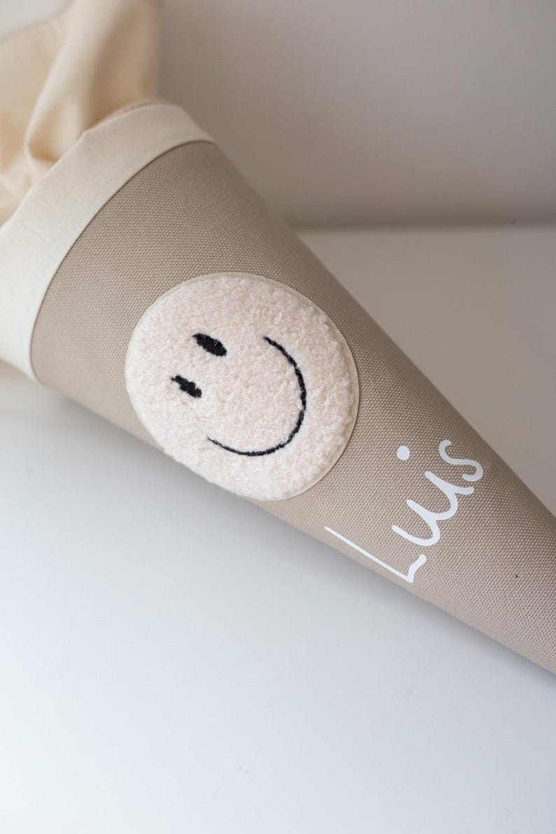 May include: A beige fabric cone-shaped gift bag featuring a circular cream-coloured fuzzy patch with a black embroidered smiley face. The name Luis is printed in white script lettering on the lower side of the cone, which has a cream fabric top.