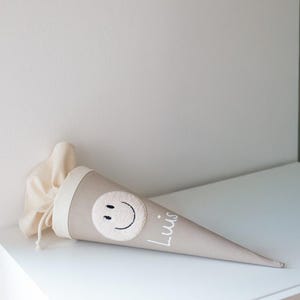 May include: A beige cone-shaped fabric gift bag featuring a cream-coloured circular patch with a black embroidered smiley face and the name Luis written in white script. The top of the bag has a cream fabric closure with a drawstring cord.