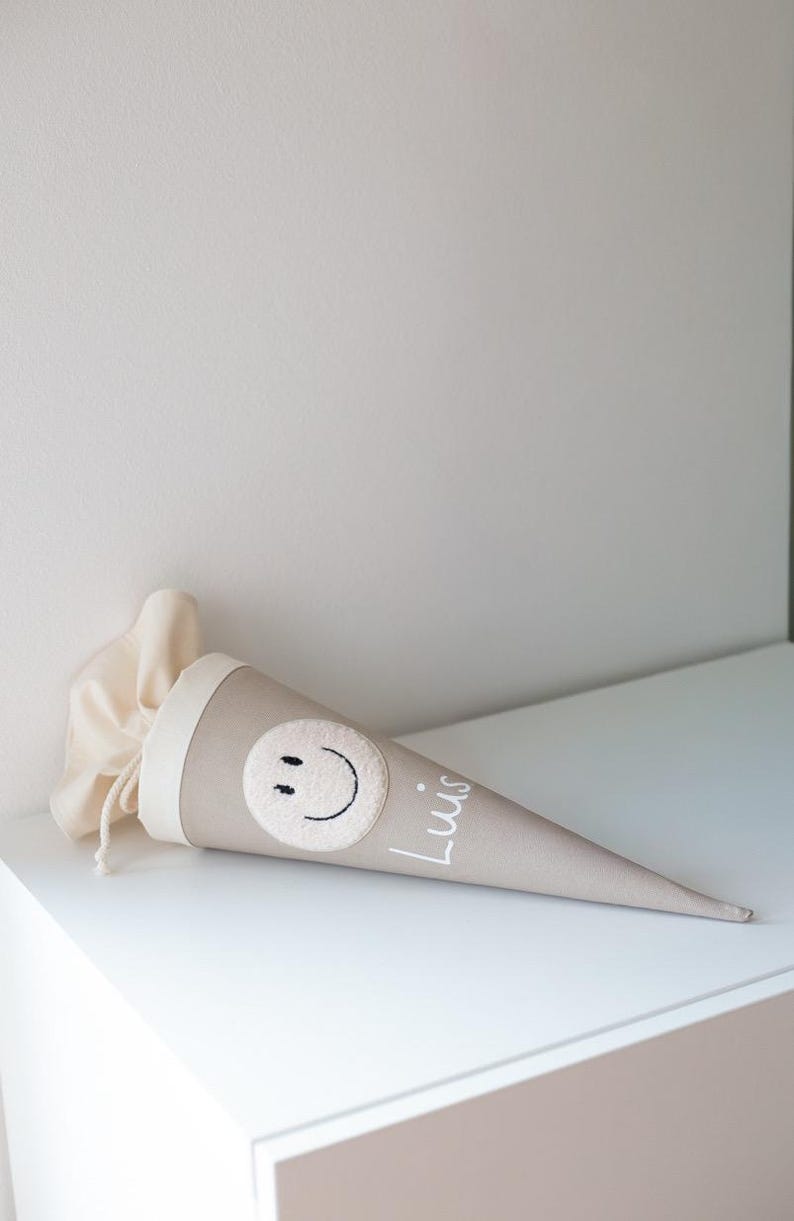 May include: A beige cone-shaped fabric gift bag featuring a cream-coloured circular patch with a black embroidered smiley face and the name Luis written in white script. The top of the bag has a cream fabric closure with a drawstring cord.
