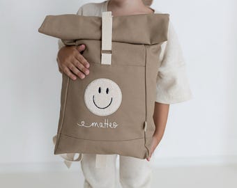 May include: A taupe roll-top backpack featuring a cream-coloured vertical strap with a metal buckle. The front displays a circular patch with an embroidered smiley face and the name "matteo" stitched in white cursive script below the patch.