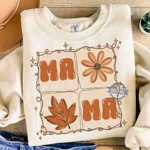 Könnte beinhalten: Ein cremefarbenes Sweatshirt mit Rundhalsausschnitt und einem Grafikdesign, das durch eine braune Linie in vier Quadrate unterteilt ist. Das Design enthält den Schriftzug "MAMA" in orangefarbenen Buchstaben, eine braune Blume und ein braunes Ahornblatt.