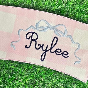 Puede incluir: Funda para taza de café de tela vichy rosa y blanca con un lazo de cinta azul y el nombre "Rylee" bordado en negro.