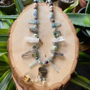 Può includere: Una lunga collana multifilo con una varietà di perline e cristalli. La collana presenta un mix di perline blu, verdi, bianche e marroni, oltre a diversi ciondoli di cristallo. La collana è rifinita con una catena dorata e un ciondolo marrone.