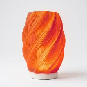 May include: An orange, spiral-shaped lamp with a white base. The lamp has a textured surface with vertical lines that create a swirling pattern. The lamp is lit from within, casting a warm glow.