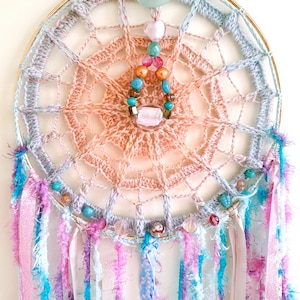 May include: A crocheted dream catcher with a pink and blue ombre design. It has a wooden bead and turquoise gemstone hanging from the centre. The dream catcher is decorated with colourful ribbons and beads.