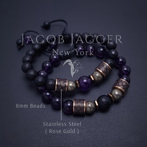 May include: Two bracelets with 8mm beads. One bracelet has black beads and the other has amethyst beads. Both bracelets have rose gold stainless steel spacers and pyrite beads. The bracelets are on a black surface. The text "JACOB JAGGER New York" is visible in the image.