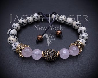 May include: A beaded bracelet featuring dalmatian jasper beads, light pink translucent beads, and a central black bead encrusted with small black stones. The bracelet includes gold-toned crown-shaped spacers and two brown end beads on black cords.