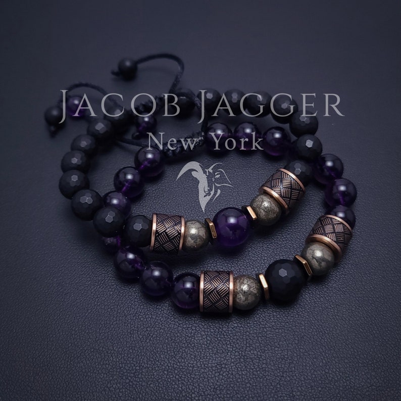 May include: Two bracelets with black and purple beads, and gold accents. The bracelets are on a dark gray surface. The text "JACOB JAGGER New York" is visible in the image.