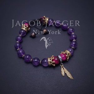 May include: A purple amethyst beaded bracelet with a gold crown charm and a tiger's eye bead. The bracelet also features a gold feather charm and the text "JACOB JAGGER New York".