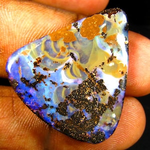 May include: A vibrant, triangular opal gemstone with a mix of colors. The stone features a base of blue and yellow hues, with areas of brown and black speckling. The opal is held in a hand, showcasing its natural beauty.