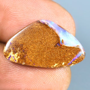 Peut inclure: Une pierre précieuse d'opale brune et blanche avec des reflets irisés violets et bleus. L'opale est de forme en larme et a une texture rugueuse et naturelle.