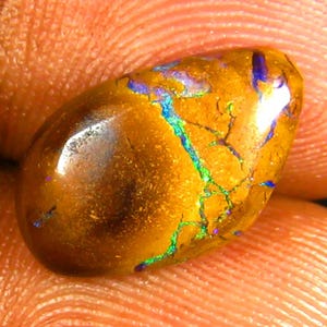 May include: A polished, oval-shaped opal gemstone. The opal has a warm, golden-brown base color with vibrant flashes of blue, green, and purple. The surface displays a network of fine cracks, adding to its unique appearance. The gemstone is held in a hand.