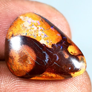 Könnte beinhalten: Ein tropfenförmiger Opal-Edelstein mit einem braunen, orangefarbenen und violetten irisierenden Schimmer. Der Opal hat eine raue, natürliche Oberfläche.