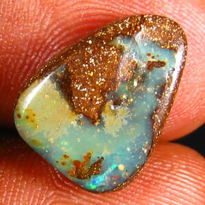 May include: A polished opal gemstone with a mix of colors. The opal features a brown matrix with white speckles, surrounding a central area of blue-green and yellow hues. The stone has a smooth, glossy surface.