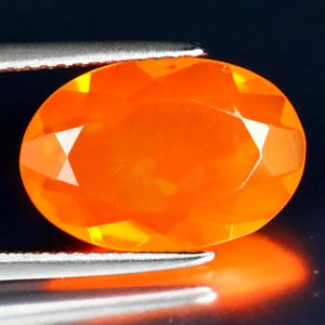 Könnte beinhalten: Ein oval geschliffener Edelstein in leuchtendem Orange, gehalten von einer silbernen Pinzette. Die Facetten des Steins reflektieren das Licht und erzeugen helle, weiße Glanzlichter. Dunkler, reflektierender Hintergrund.