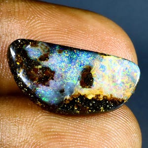 May include: A polished, oval-shaped opal gemstone. The opal displays a vibrant play-of-color with blues, greens, and yellows, set against a dark, speckled matrix. The gemstone is held between fingers.