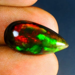 May include: A teardrop-shaped black opal gemstone with iridescent green and red flashes. The opal is held in a hand.