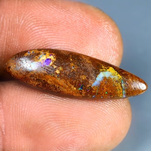 Könnte beinhalten: Ein länglicher, polierter Opal-Edelstein mit einer satten braunen Grundfarbe. Der Opal weist schillernde Blitze in Blau, Lila und Grün auf, die einen auffälligen visuellen Kontrast erzeugen. Der Stein wird zwischen Fingern gehalten, was seine natürliche Schönheit zeigt.