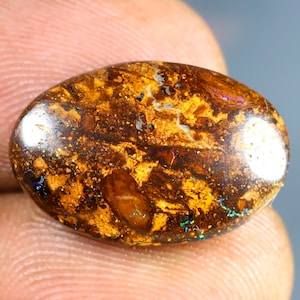 Peut inclure: Une opale ovale de couleur brune avec des reflets irisés de vert, bleu et violet. L'opale a une surface rugueuse et texturée et est taillée en cabochon.