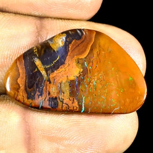 May include: A close-up of a brown, orange, and black opal gemstone with iridescent green and blue flashes. The opal is irregularly shaped and has a smooth, polished surface.