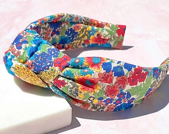 May include: A floral headband with a knotted design. The headband features a colorful pattern of red, blue, yellow, and green flowers on a cream background. The accessory is resting on a white surface against a pink backdrop.