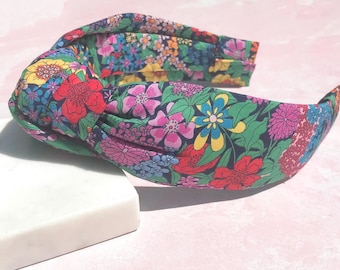 May include: A floral print headband with a knotted design. The headband features a vibrant pattern of red, pink, yellow, and blue flowers on a dark green background. The accessory is resting on a white rectangular block against a pink backdrop.