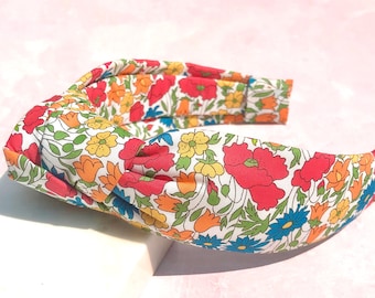 May include: A floral headband featuring a vibrant pattern of red poppies, yellow, orange, and blue flowers on a white background. The headband is made of fabric and is designed to be worn on the head. The accessory is placed on a white surface.