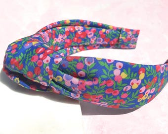 May include: A fabric headband with a knotted top. The headband features a vibrant floral pattern with red, pink, and yellow flowers on a blue background. The accessory is designed for hair styling and fashion.