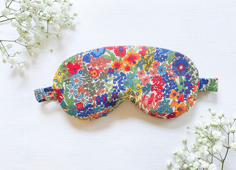 May include: A floral patterned sleep mask with a white background and colorful flowers in shades of red, blue, yellow, and green. The mask has a strap that goes around the head.