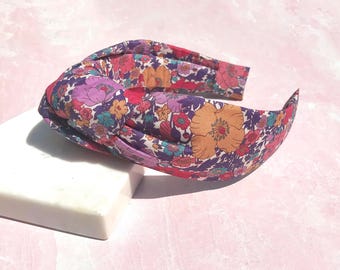 May include: A floral headband with a knotted design. The headband features a vibrant pattern of orange, purple, red, and teal flowers on a purple background. The accessory is displayed on a white surface against a pink backdrop.