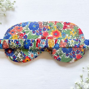 May include: A floral eye mask with a white background and a blue, red, and yellow floral pattern. The mask has an elastic band that is also covered in the same floral fabric.