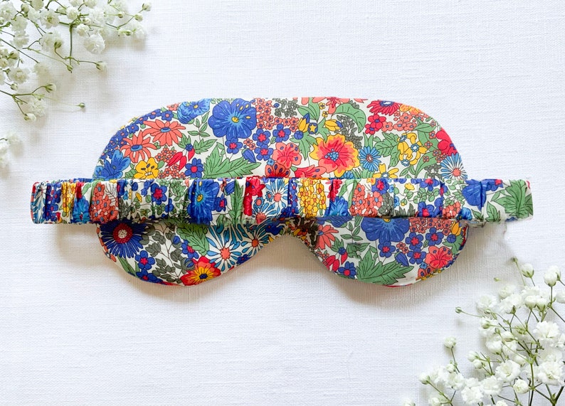 May include: A floral eye mask with a white background and a blue, red, and yellow floral pattern. The mask has an elastic band that is also covered in the same floral fabric.