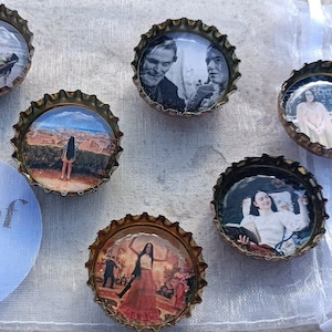May include: Six round bottle caps with images printed on them. The images include a cityscape, a woman in a dress, a woman in a white shirt, two men, and a woman in a pink dress. The bottle caps are gold colored with a silver rim.