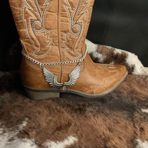 May include: A brown leather cowboy boot with decorative stitching and a silver-colored chain ankle bracelet featuring a wing charm. The boot is set against a backdrop of a cowhide rug.