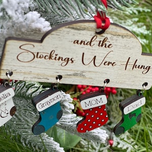May include: A wooden Christmas ornament with the text "And the Stockings Were Hung."  Four small stockings hang from the ornament, each with a different name and design: Amelia with a white stocking and a horse design, Grayson with a blue stocking and a snowflake design, Mom with a red stocking and white polka dots, and Dad with a green stocking and a reindeer design.