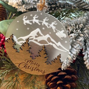 May include: A round wooden Christmas ornament with a white silhouette of a mountain range and Santa's sleigh flying over the mountains. The ornament has the text "Merry Christmas" inscribed on the bottom.
