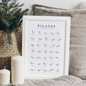 Peut inclure: Affiche Pilates encadrée de blanc, avec des dessins au trait noirs d'exercices. Le mot "PILATES" est en haut. Deux bougies blanches et un panier tressé avec de la verdure sont en arrière-plan. Un oreiller et une couverture gris sont également en arrière-plan.