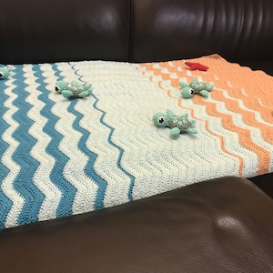 May include: A crocheted baby blanket with a blue and white wave pattern on one side and a peach and white wave pattern on the other side. Five small crocheted sea turtles are placed on the blanket.