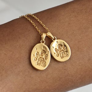 May include: A gold-toned necklace featuring two oval-shaped pendants suspended from a delicate chain. Each pendant is engraved with a textured paw print design, creating a detailed and distinct visual element on the surface of the metal charms.