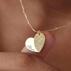 May include: A gold-colored necklace with a heart-shaped pendant. The pendant features a fingerprint design and the word "Mom" engraved on it. The necklace is a delicate piece of jewelry.