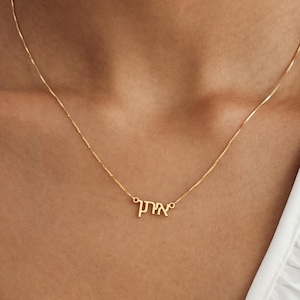 May include: A gold-toned necklace featuring a delicate chain and a central pendant displaying the Hebrew name Eitan in a script font. The metal has a polished finish, and the pendant is securely attached to the chain on both sides.