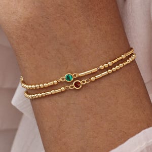May include: Two delicate gold bracelets, each with a string of small beads and a single gemstone. One bracelet features a green gemstone, the other a red gemstone. The bracelets are worn on a wrist.