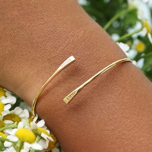 May include: A gold-toned open cuff bracelet featuring two tapered ends. The bracelet is engraved with the name BRIAN on one end and the name EMMA on the other end. The metal has a smooth, polished finish and a thin, flexible wire-like structure.