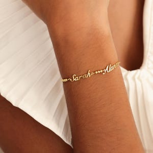 May include: A gold-colored bracelet with the names "Sarah" and "Alan" in cursive script. The bracelet is worn on a person's arm, with a white background.